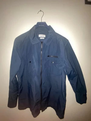 Blå tunn jacka från Strömberg  - Snygg marinblå overshirt från strömberg med klassisk krage, dragkedja framtill och praktiska fickor med knappar och zip. Jackan är tillverkad i lätt polyester och har långärmad passform, perfekt för lager på lager.