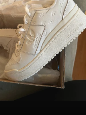 Adidas Forum Bold W vita sneakers - Säljer ett par Adidas Forum Bold W sneakers i helvitt skinn med präglade Adidas-loggor och chunky plattformssula. Skorna har snörning, rund tå och diskreta detaljer som ger en clean och modern vibe. Perfekta för dig som gillar stilrena och trendiga sneakers.