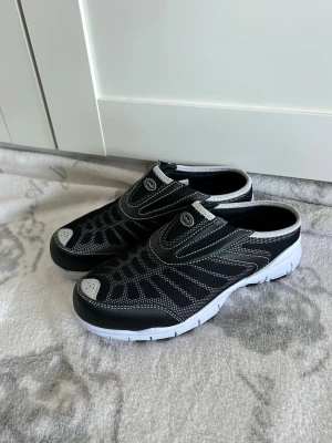 Svarta slip-on sneakers med vit sula - Svarta slip-on sneakers från Scholl med vit sula och grå detaljer. Ovandelen är i mesh och syntet med kontrasterande sömmar och rund tå. Skorna har en sportig design och är lätta att ta på och av, perfekta för dig som gillar bekväma och stilrena skor.