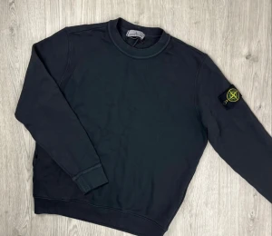 Svart Stone Island sweatshirt - Svart sweatshirt från Stone Island med klassisk rund hals och ikonisk logotyp-patch på vänster ärm. Tröjan är i mjuk bomull och har ribbade muddar vid ärmslut och nederkant. Perfekt för dig som gillar stilren streetwear med premiumkänsla.