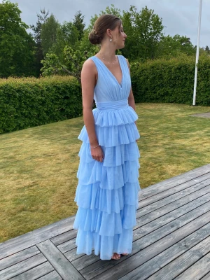 Ljusblå balklänningmed volanger - Säljer en ljusblå balklänning med flera lager volanger och v-ringad omlott upptill. Klänningen är ärmlös och har ett brett midjeband som markerar midjan snyggt. Perfekt för bal eller student! Köpt på bubbelrom. Inga defekter