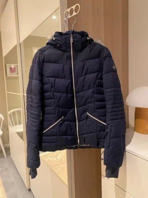 Jacka - Snygg mörkblå jacka från McKinley med quiltad design, huva med grå fuskpäls och två dragkedjeförsedda fickor. Jackan har lång ärm, hög krage och silverfärgade detaljer. Perfekt för kalla dagar och har en modern, sportig look. Kan användas som skidjacka. Storlek 38. 