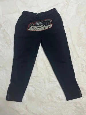 Svarta Trapstar shooters mjukisbyxor med röd söm - Svarta mjukisbyxor från Trapstar med stort broderat 'London Shooters' motiv och panter på framsidan. Byxorna har snörning i midjan och dragkedjor vid bensluten. Materialet är mjukt och bekvämt. Storleken är L men passar även för S-M då dem är ganska så tajta. Har även Hoodie till och försäljning på den kan diskuteras om man vill ha hela settet! Nästan helt oanvända! 