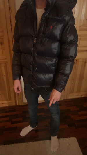 Svart pufferjacka från Polo Ralph Lauren - Säljer en svart puffer från Polo Ralph Lauren med röd logga på bröstet. Jackan är pösig och har huva, dragkedja framtill och en quiltad design. Perfekt för kalla dagar och riktigt snygg till jeans. Säljer pga att den va lite för stor och vill få tbx lite pengar. Storlek S men den va rätt stor och passar M. Modellen är 180cm
