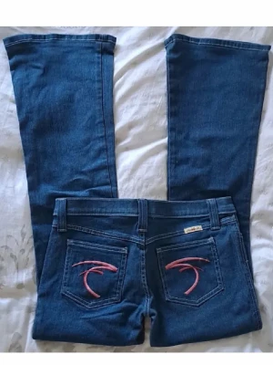 Blå bootcut jeans från Frankie B. - Snygga blå bootcut jeans från Frankie B. med rosa broderi på bakfickorna. Jeansen har klassisk femficksdesign och är tillverkade i denim med stretch för extra komfort. Perfekta för dig som gillar en retroinspirerad look med unika detaljer.