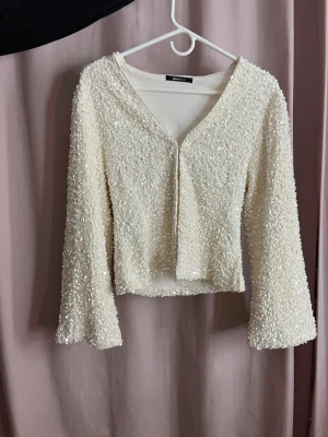 Vit paljettopp från Gina Tricot - Supersnygg vit topp från Gina Tricot täckt med glittrande paljetter. Modellen är v-ringning och långa ärmar. Perfekt för dig som vill sticka ut lite extra. Toppen har en mjuk insida och känns bekväm mot huden.