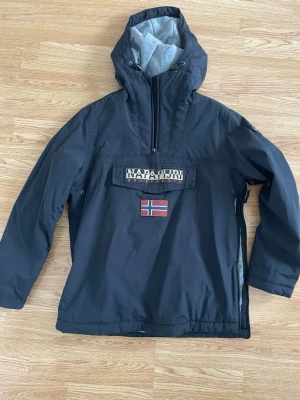 Napapijri svart parkas med fleece - Svart parkasjacka från Napapijri med stor huva, halv dragkedja och grått fleecefoder. Stor magficka med Napapijri-logga och norsk flagga på bröstet. Dragkedja i sidan för smidig på- och avtagning. Materialet är slitstarkt och vindtätt.