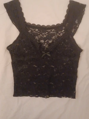 Svart spetstopp med rosett - Snygg svart topp i spets med breda axelband och söt liten rosett framtill. Djup v-ringning och genombrutet spetsmönster ger en feminin look. Perfekt att styla under en skjorta eller kavaj.Vet ej märket för köpte på vinted och det står ingen lapp på plagget. Kolla sista bilden för defekter. 