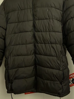 Svart Jacka - Säljer en svart pufferjacka med huva och dragkedja framtill. Jackan har två fickor med dragkedja och breda, horisontella kanaler som ger en klassisk pufferlook. Perfekt för kalla vinterdagar och riktigt skön att bära.