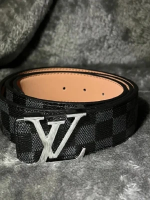 Svart rutigt  bälte i skinn - Snyggt svart bälte med klassiskt rutigt mönster och stor logga i silver på spännet. Bältet är gjort i skinn och har en stilren design som passar till många olika outfits.