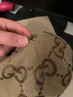 Beige GG-keps från Gucci - Säljer en beige keps från Gucci med det klassiska GG-mönstret i brunt över hela kepsen. Kepsen har böjd skärm och bruna detaljer längs kanten. Tillverkad i ett slitstarkt tygmaterial som ger en exklusiv känsla. Den e gammal använd länge och smutsig men den är äkta 