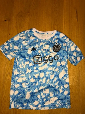 Ajax blå och vit träningströja - Säljer en Ajax träningströja från Adidas i blått och vitt kamouflagemönster. Tröjan har korta ärmar, rund hals och Ajax-logga på bröstet samt Ziggo-tryck framtill. Perfekt för fotbollsträning eller sportiga dagar.
