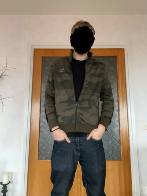 Camo ziptröja från Abercrombie & Fitch - Snygg ziptröja i camomönster från Abercrombie & Fitch. Tröjan har hög krage, dragkedja framtill och en patch med logga på bröstet. Perfekt för en avslappnad streetstil. Materialet är mjukt och bekvämt, passar till jeans eller joggers.