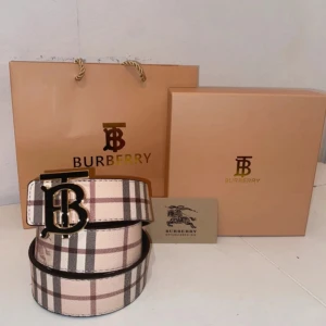 Burberry rutiga skärp med B-spänne - exklusiva Burberry-bälten med ikoniskt rutmönster och stort B-spänne i guld och svart. Ett bälte i beige, brunt och rött, det andra i svart och grått. Båda är tillverkade i skinn och har klassisk Burberry-logga. Perfekt accessoar för att lyfta din outfit.
