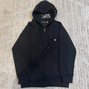 Svart hoodie från Polo Ralph Lauren - Klassisk svart hoodie med dragkedja från Polo Ralph Lauren. Tröjan har huva med dragsko, två fickor framtill och den ikoniska vita logon broderad på bröstet. Perfekt för en avslappnad och stilren look.