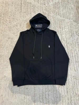 Svart hoodie från Polo Ralph Lauren - Klassisk svart hoodie från Polo Ralph Lauren med huva och dragsnören. Har en vit broderad logga på bröstet och en stor magficka framtill. Tillverkad i mjukt bomullsmaterial och har ribbade muddar vid ärmslut och nederkant.