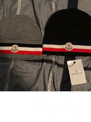 Moncler mössa med randigt band - En Moncler mössor, en i grått och en i svart, båda med ett randigt band i svart, vitt och rött samt Moncler-logga framtill. Klassisk och stilren design som passar perfekt till vintern. Mössorna är ribbstickade och har en mjuk känsla.