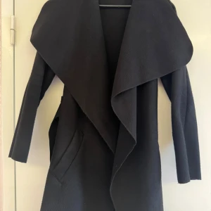 Svart omlott kappa med bälte - Snygg svart kappa med bred krage och omlott-design. Jackan har ett matchande bälte i midjan och två fickor framtill. Långärmad och med en stilren, loose passform som ger en chill vibe. Har endast ett få stygn som gått uppp under den högra armen, men lätt att sys ihop igen 😊