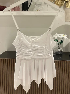Vit volang linnetopp med smala band - Säljer en vit linnetopp med smala axelband och snygga volangdetaljer nedtill. Toppen har rynkade detaljer framtill och en flowy, asymmetrisk nederkant som ger en riktigt unik look. Stretchigt material. Perfekt för dig som vill ha något extra till outfiten. Står värken märke eller storlek men skulle säga M. Axelbredd: 23cm. Längd: 59cm. Byst: 