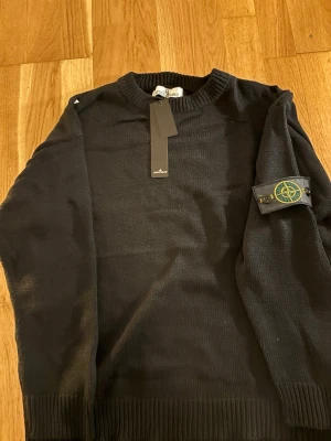 Svart stickad tröja från Stone Island - Svart stickad tröja från Stone Island med klassisk rund hals och ikonisk patch på vänster ärm. Tröjan har ribbade muddar och är långärmad, perfekt för dig som gillar stilrena och tidlösa plagg med streetwear-vibe.