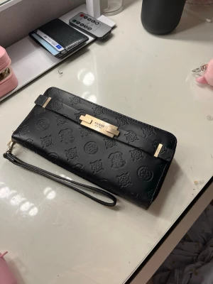 Svart plånbok från Guess med mönster - Snygg svart plånbok från Guess i konstläder med präglat logomönster och guldfärgade detaljer. Plånboken har handledsrem, dragkedjefack på baksidan och metallspänne framtill med Guess-logga. Perfekt för att hålla koll på kort och cash.
