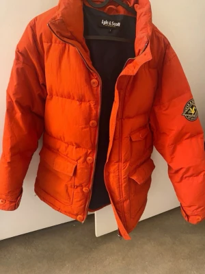 Röd herr dunjacka från Lyle & Scott S  - Snygg röd dunjacka från Lyle & Scott med hög krage, dragkedja och knappar framtill. Jackan har två stora fickor med lock och ett tydligt Lyle & Scott-märke på ena ärmen. Perfekt för kalla vinterdagar.