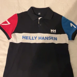 Flerfärgad piké från Helly Hansen - Snygg pikétröja från Helly Hansen med svart bas, vit rand över bröstet och blått tryck. Ena ärmen är röd och den andra blå, båda med vita detaljer. Klassisk krage och knappar framtill. Perfekt för en sportig och avslappnad look.