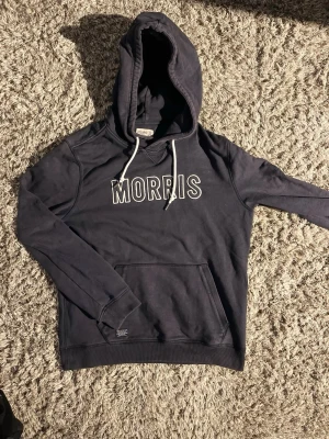 Mörkblå hoodie från Morris🙌🤩 - Snygg mörkblå hoodie från Morris med vit logotyp på bröstet och vita dragsnören i huvan. Klassisk känguruficka framtill och ribbade muddar vid ärmar och nederkant. Perfekt för en avslappnad och stilren look. 