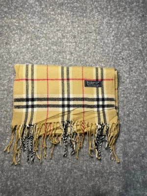 Burberry Halsduk Beige - Klassisk Burberry-halsduk i 100 % lammull. Mjuk och varm med tidlöst rutmönster i beige, svart, vitt och rött. Originaletikett, Made in England. 2B
