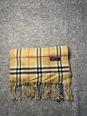 Burberry Halsduk Beige - Klassisk Burberry-halsduk i 100 % lammull. Mjuk och varm med tidlöst rutmönster i beige, svart, vitt och rött. Originaletikett, Made in England. 1B