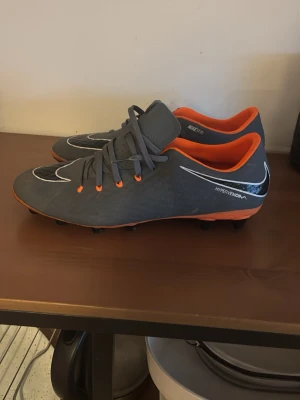 Nike Hypervenom grå fotbollsskor - Snygga grå Nike Hypervenom fotbollsskor med orange detaljer och svart sula. Skorna har en låg profil, snörning och innersula i orange. Tillverkade i syntetmaterial med NIKESKIN för extra känsla och kontroll på planen. Perfekta för dig som vill sticka ut på matchen.