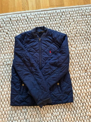 Marinblå quiltad vindjacka Polo Ralph Lauren - Snygg marinblå vindjacka från Polo Ralph Lauren med quiltat mönster, dragkedja framtill och två fickor med tryckknappar. Jackan har en liten röd logga broderad på bröstet och ribbade muddar vid ärmsluten. Perfekt för en stilren och sportig look.