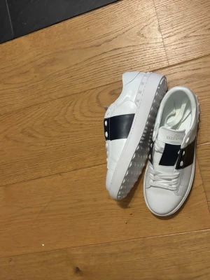 Vita Valentino sneakers med svart band - Snygga vita sneakers från Valentino med ett brett svart band över sidan och coola nitar. Skorna har en chunky sula med rutmönster undertill och klassisk snörning. Tillverkade i läder för en lyxig känsla och stilren look.