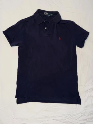 Mörkblå pikétröja från Ralph Lauren - Klassisk mörkblå pikétröja från Ralph Lauren med korta ärmar och två knappar vid kragen. Tröjan har den ikoniska röda logotypen broderad på bröstet och är tillverkad i mjuk bomull. Perfekt för en clean och stilren look.