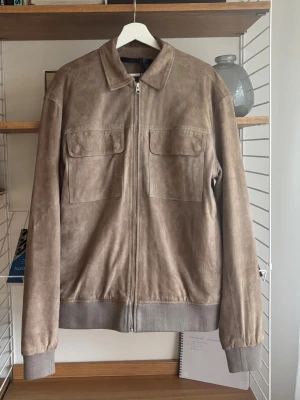 Mockajacka från Tiger Of Sweden  - Stilren beige mockajacka med dragkedja framtill och två stora bröstfickor med lock. Jackan har ribbade muddar vid ärmslut och midja samt klassisk krage. Perfekt nu till våren. Storlek M. Ord pris 5499