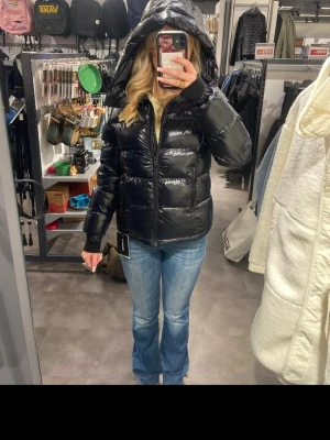 Svart glansig dunjacka med huva - Svart puffer dunjacka med glansig finish och huva. Jackan har dragkedja framtill och två sidofickor. Perfekt för kyliga dagar och ger en modern look till din outfit.