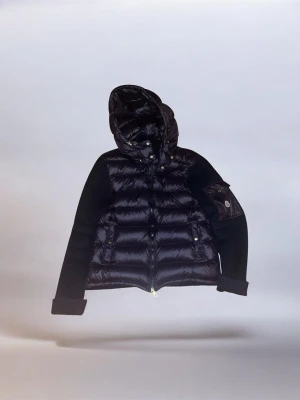 Svart dunjacka med huva från Moncler - Snygg svart dunjacka från Moncler med quiltad kropp och stickade ärmar. Jackan har en stor huva, dragkedja framtill och fickor med knappdetaljer. Perfekt för kalla dagar och ger en cool streetwear-look.