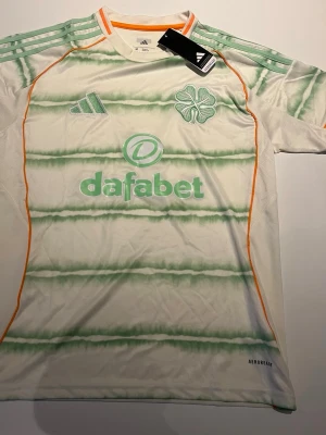 Celtic bortatröja Adidas 2023/24 - Vit Celtic bortatröja från Adidas med gröna horisontella ränder, orange detaljer och klubbmärke på bröstet. Tröjan har Aeroready-material för bra ventilation och sponsorlogga på framsidan. Perfekt för fotbollsträning eller match.