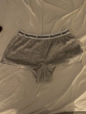 Velourshorts Juicy Couture - Mysiga grå och lila velourshorts från Juicy Couture med elastisk midja och logotyp i svart på det vita midjebandet. Baksidan på de gråa finns texten 'Juicy' i glittrande stenar. Perfekta för chill hemma eller som loungewear.