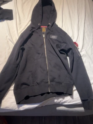Svart zip hoodie Alpha Industries - Säljer en svart hoodie från Alpha Industries med dragkedja framtill och stor vit logga på ryggen. Hoodien har huva, en ficka på armen, två stora fickor fram och ribbade muddar. Perfekt för en avslappnad och cool streetwear-look.