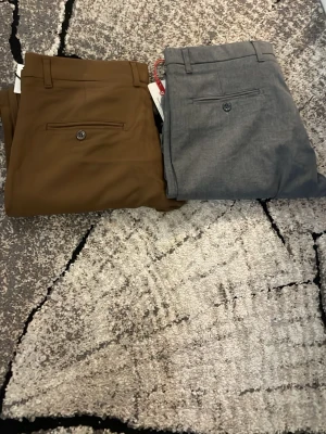 Bruna och grå kostymbyxor herr - Två par stilrena kostymbyxor i klassisk rak modell, ett par i brun och ett par i grå färg. Byxorna har hällor för bälte och bakfickor med knapp. Perfekta för en dressad look. Materialet ser ut att vara en blandning av polyester och bomull.