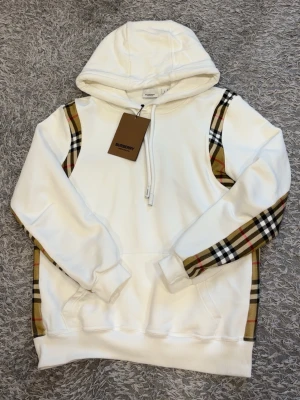 Burberry hoodie - Snygg vit hoodie från Burberry med klassiska beige, svarta och röda rutiga detaljer längs ärmar och axlar. Hoodien har huva med dragsko och känguruficka framtill. Tillverkad i mjukt bomullsmaterial och har ribbade muddar vid ärmslut och nederkant.