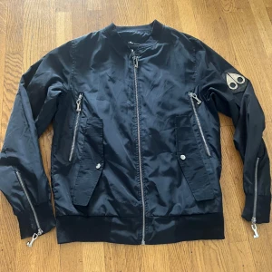 Svart bomberjacka Moose Knuckles - Snygg svart bomberjacka från Moose Knuckles med ikonisk metallogo på ärmen och broderad design på ryggen. Jackan har flera dragkedjor, ribbade muddar och klassisk passform. Tillverkad i glansig polyester som ger en clean och modern look.