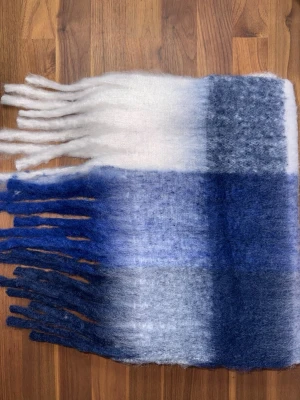 Rutig halsduk i blått och vitt - Mjuk och fluffig halsduk med rutigt mönster i blått och vitt. Halsduken har långa fransar i båda ändar och är perfekt för att hålla värmen under kalla dagar. Materialet känns ulligt och ger en mysig känsla. Halsduken liknar ”Acne Studios halsdukarna och är oanvänd (ny).