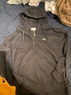 Svart hoodie från Lacoste - Svart zip-hoodie från Lacoste med klassisk krokodillogga på bröstet. Modellen har huva med dragsko, två fickor framtill och ribbade muddar. Perfekt för en chill och stilren look.storlek är xl men skulle säga den passar L ochså 