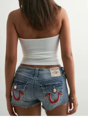 True Religion Joey Cut-Off jeansshorts - Säljer ett par grymma blå jeansshorts från True Religion, modell Joey Cut-Off. De är lågmidjade med råa, fransiga kanter och har röda detaljer på bakfickorna. Klassisk femficksdesign och coola kopparfärgade knappar. Perfekta för sommaren! Har bara använt ett fåtal gånger 