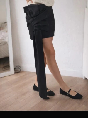 Svart minikjol med stor rosett - Trendig svart minikjol med unik stor rosettdetalj och lång hängande tygdel framtill. Kjolen har en figurnära passform och är tillverkad i ett slätt tyg som ger en stilren look. Perfekt för dig som vill sticka ut med en cool och modern twist. PRISLAPP KVAR!!