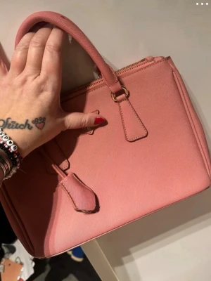 Rosa handväska med gulddetaljer - Snygg rosa handväska i fyrkantig modell med två rejäla handtag och guldfärgade dragkedjor. Väskan har flera fack och är tillverkad i ett slätt, konstläderliknande material. Perfekt för dig som vill ha en stilren och trendig accessoar. Klister på handtagen som jag inte vet hur man får bort. 