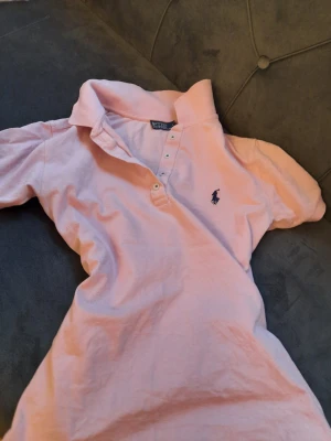 Ljusrosa pikétröja från Polo Ralph Lauren - Klassisk ljusrosa pikétröja från Polo Ralph Lauren med korta ärmar och blå broderad logga på bröstet. Tröjan har krage och knappar framtill. Perfekt för en avslappnad men ändå stilsäker look.(Det är en doupe fast ser exakt likdan ut både med kvalite och logga)🤍