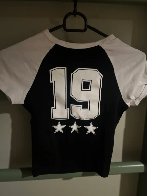 Svartvit t-shirt med tryck och stjärnor - Cool svart t-shirt med vita ärmar och stort tryck med siffran 19 och tre stjärnor på ryggen. Klassisk basebollstil med kontrastfärgade ärmar. Perfekt för en sportig look.